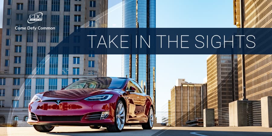A red sports car parked at the top of a parking garage with beautiful, tall corporate buldings in the background. Across the top, the words: take in the sights.