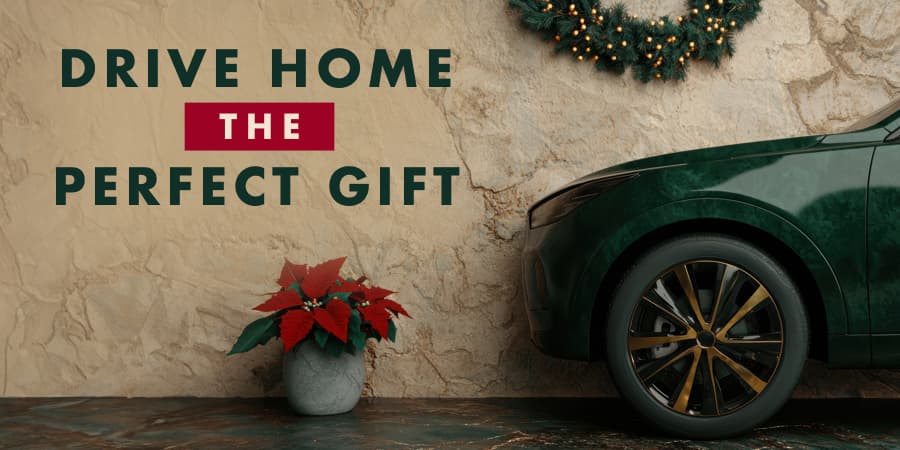 A poinsetta plant in front of the side front profile of a car parked against a textured beige wall. At the left, the words: drive home the perfect gift.