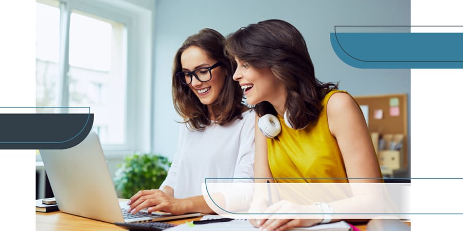 Two young women sitting at a desk looking at an open laptop.