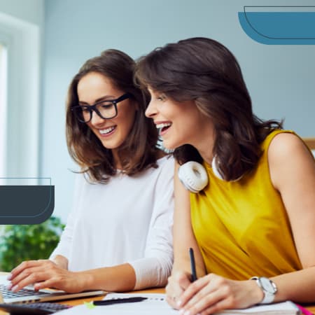 Two young women sitting at a desk looking at an open laptop.