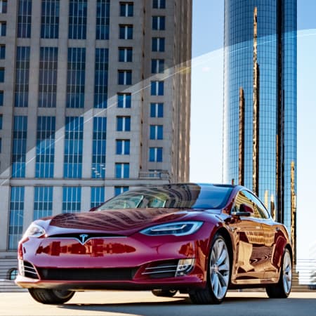 A red sports car parked at the top of a parking garage with beautiful, tall corporate buldings in the background.
