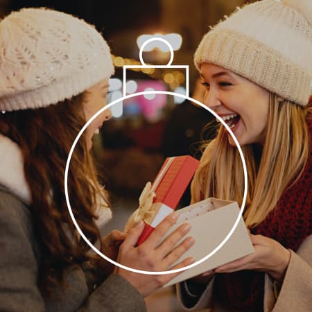 Two young adults in winter attire smiling while one presents a gift to the other.
