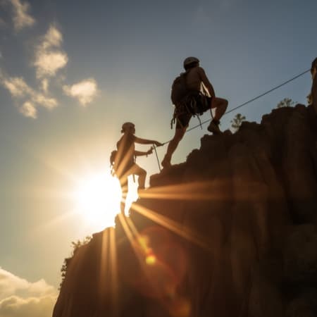 Two hikers climbing a mountain with the sun and sky in the background.