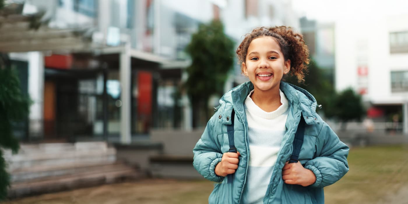 A young student wearing a backpack smiling while standing in front of a school entrance.