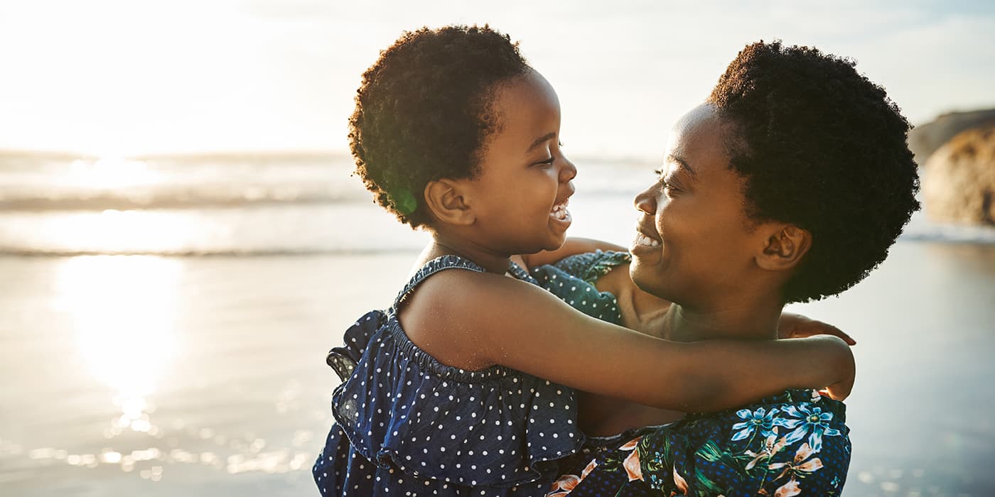 A mother carrying her young daugther while standing on the ocean shoreline.