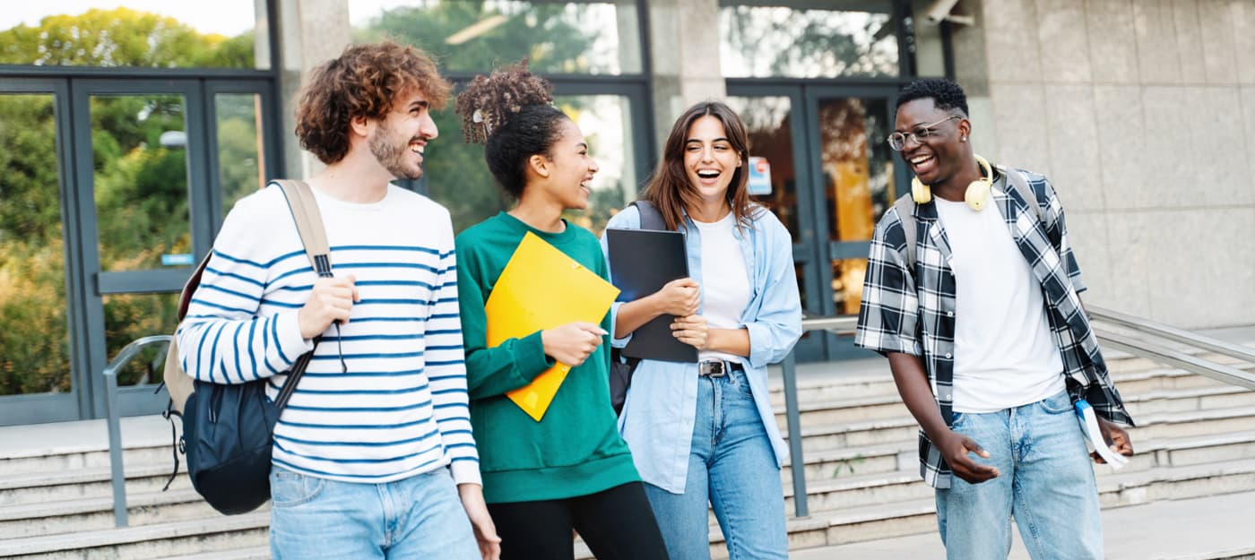 Four college students walking and talking together outside a campus building while carrying backpacks, notebooks, and school materials.