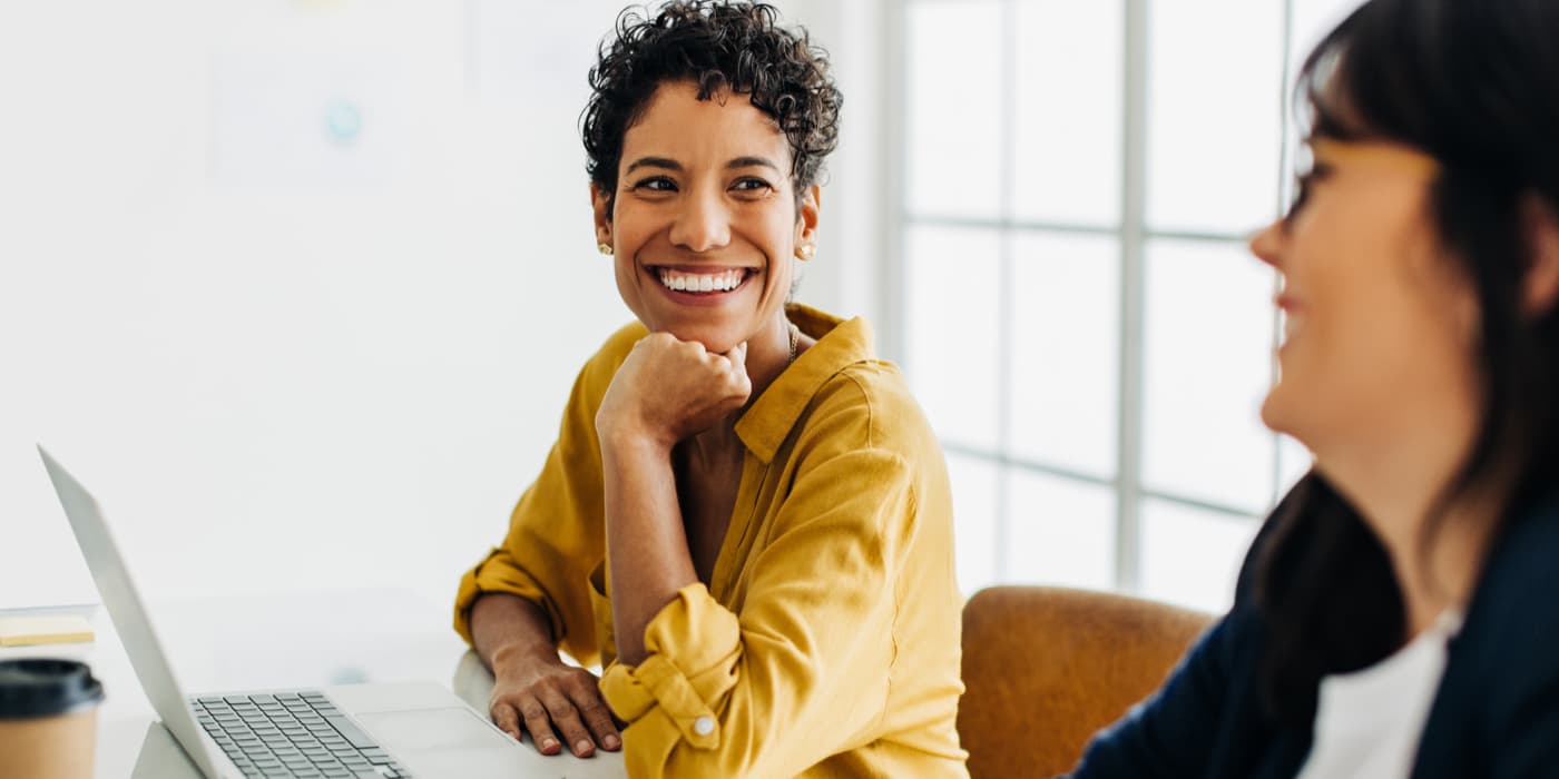A smiling woman sitting at her desk looking at another woman beside her.