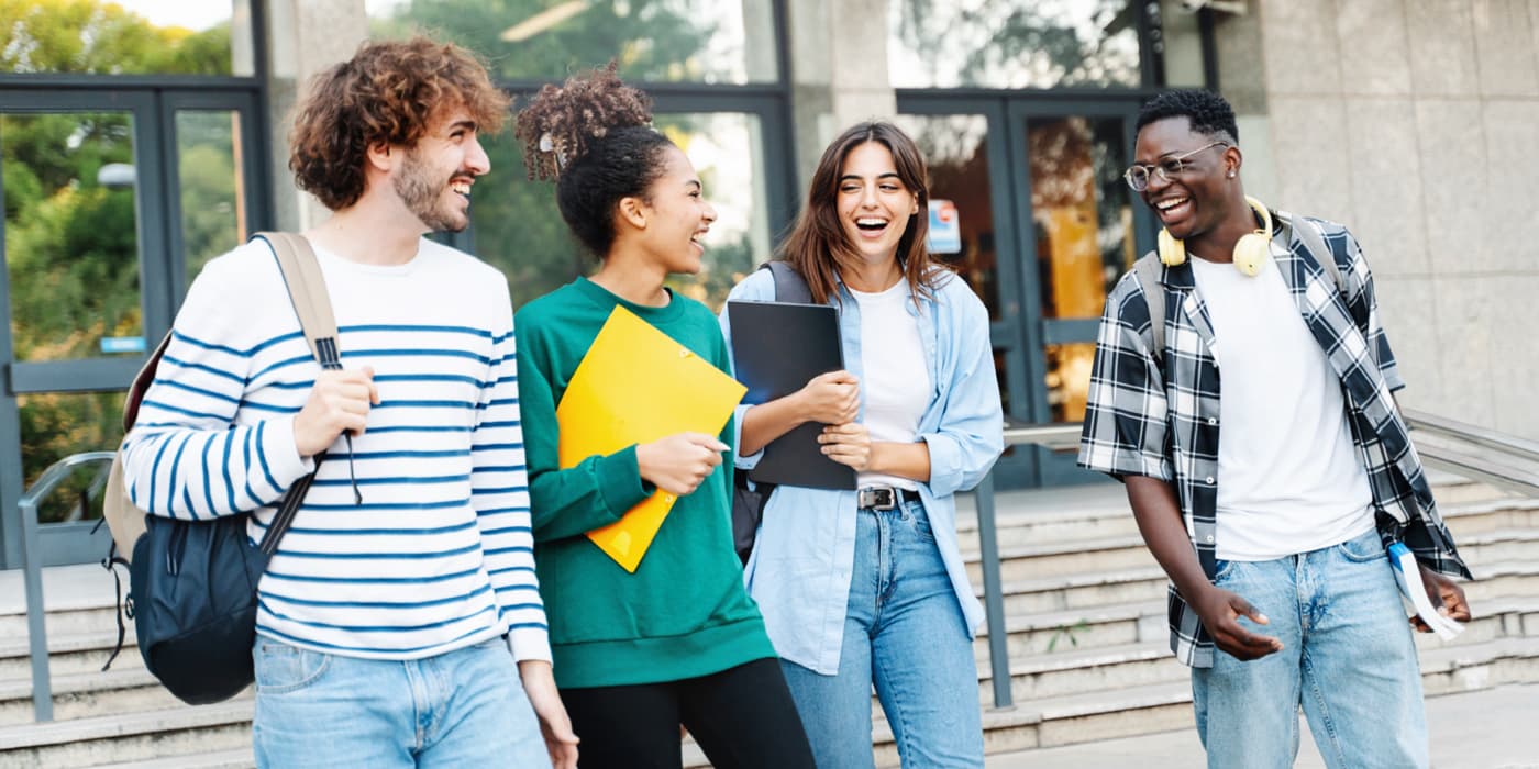 Four college students walking and talking together outside a campus building while carrying backpacks, notebooks, and school materials.