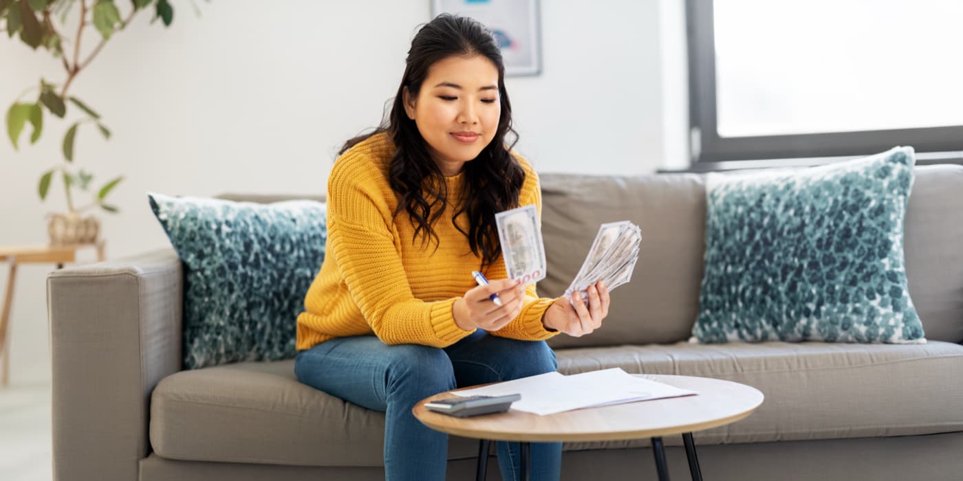 Woman sitting on a couch at home reviewing and counting cash with bills and a calculator on the table in front of her.