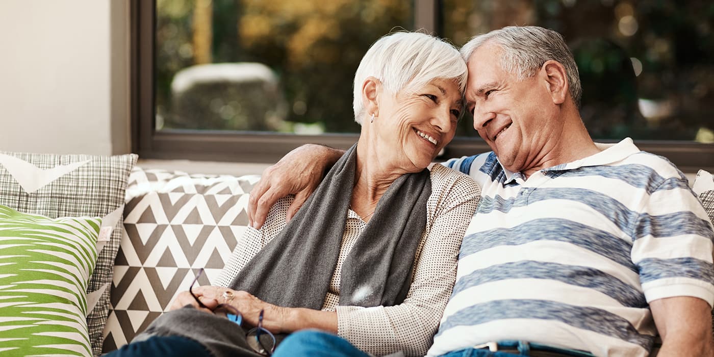 An elderly couple sitting on a couch with their heads tilted together.